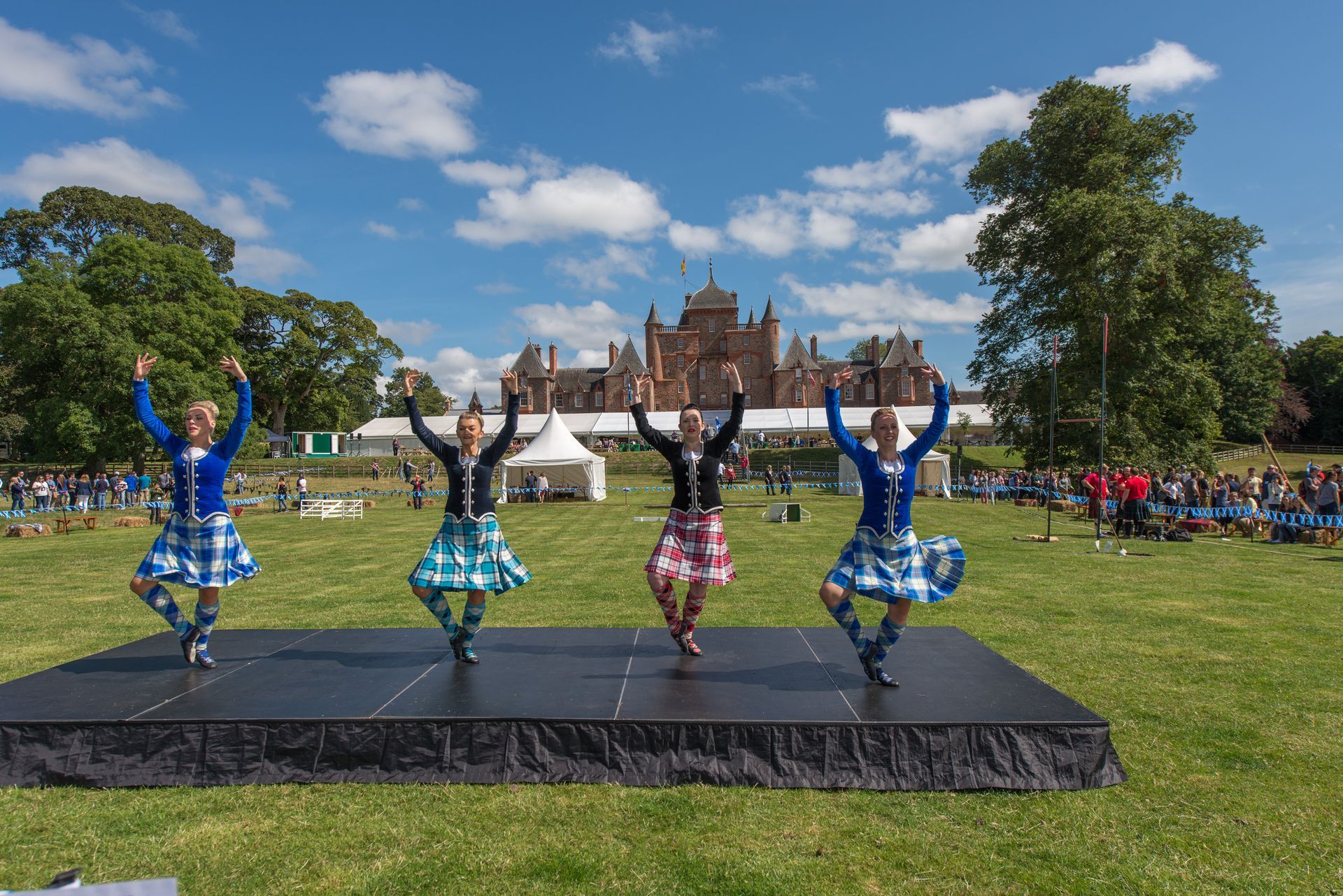 Four dancers in tartan kilts on a stage, arms raised, performing outdoors. A building is in the background.