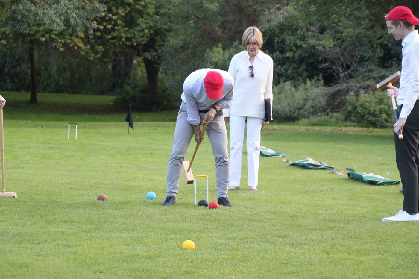 People playing croquet on a green lawn. One in a red hat swings a mallet. Others watch.