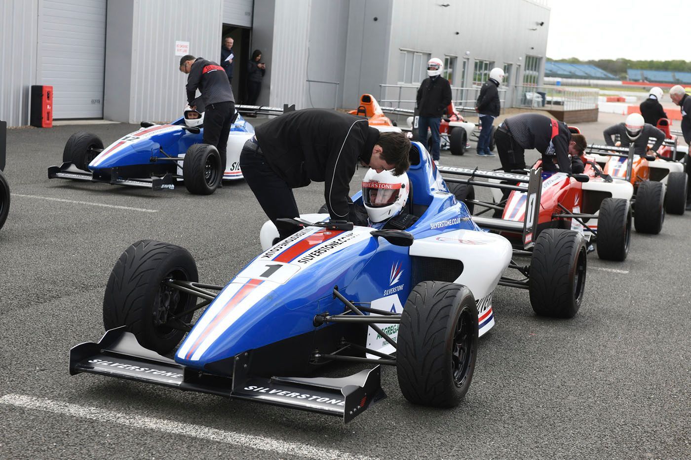Formula race cars lined up, drivers and crew working on them at a racetrack. Cars are blue, white, and red.