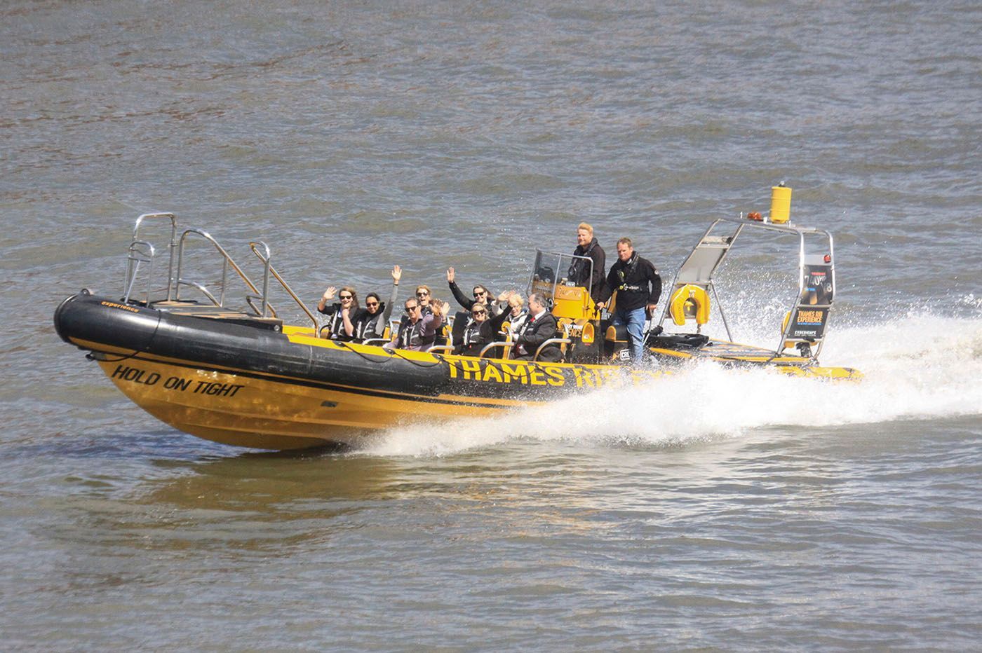 Yellow and black Thames Rockets boat with passengers on the Thames River, London. People wave, boat speeds.