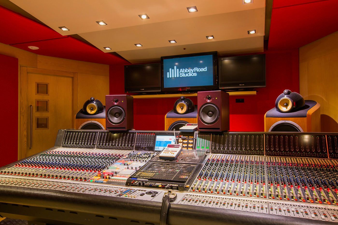 Recording studio control room with mixing console and speakers. Red and beige walls, screens.
