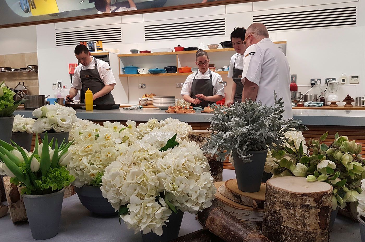 Chefs prepare food in a kitchen, surrounded by white flowers.