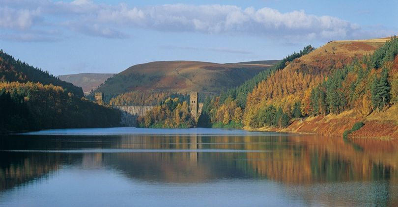 Serene lake reflecting autumn hills and trees, with a dam in the distance.