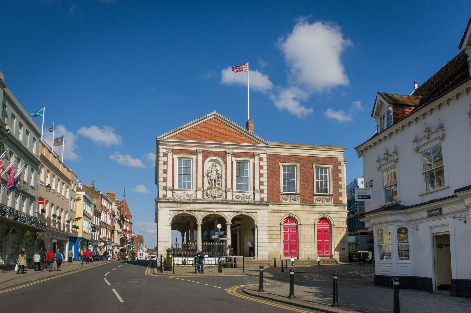 Market square in front of a historic building with red doors, under a blue sky, street with some people.