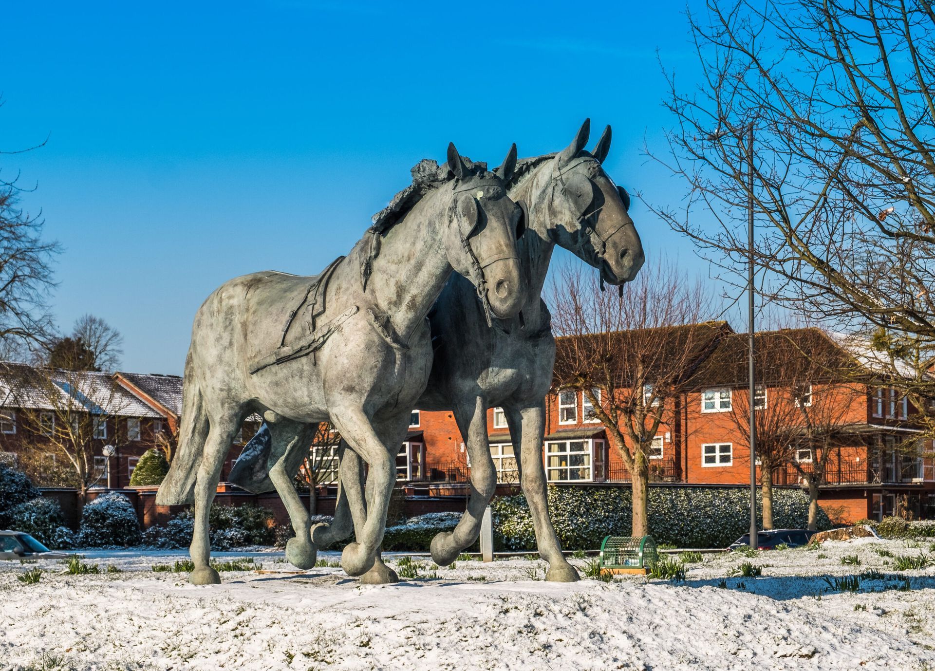 Bronze sculpture of two horses walking in a snowy park, houses in the background.