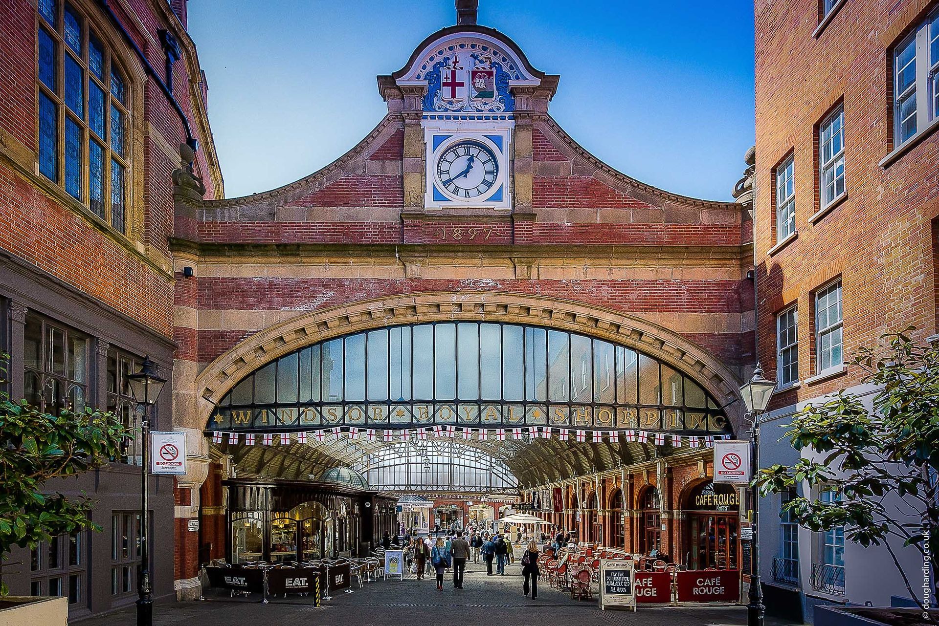 Red brick archway with clock over a bustling shopping area; trees line the side.