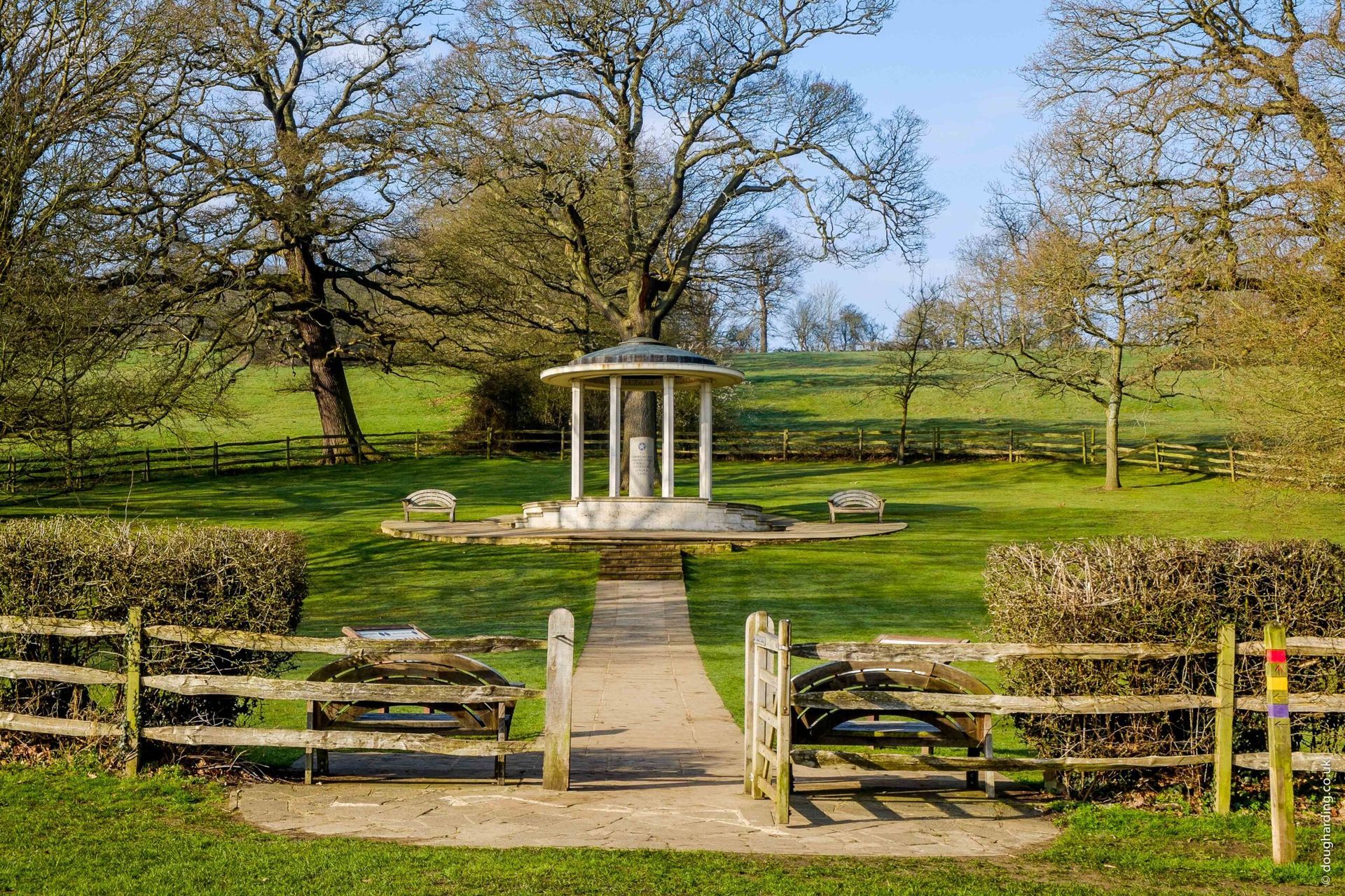 Park scene with a white gazebo, pathway, and wooden fence. Trees and green grass surround it.