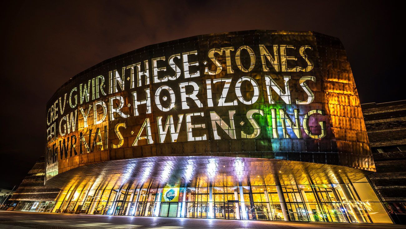 Night view of the Wales Millennium Centre in Cardiff, illuminated with projected Welsh text, in shades of gold and green.
