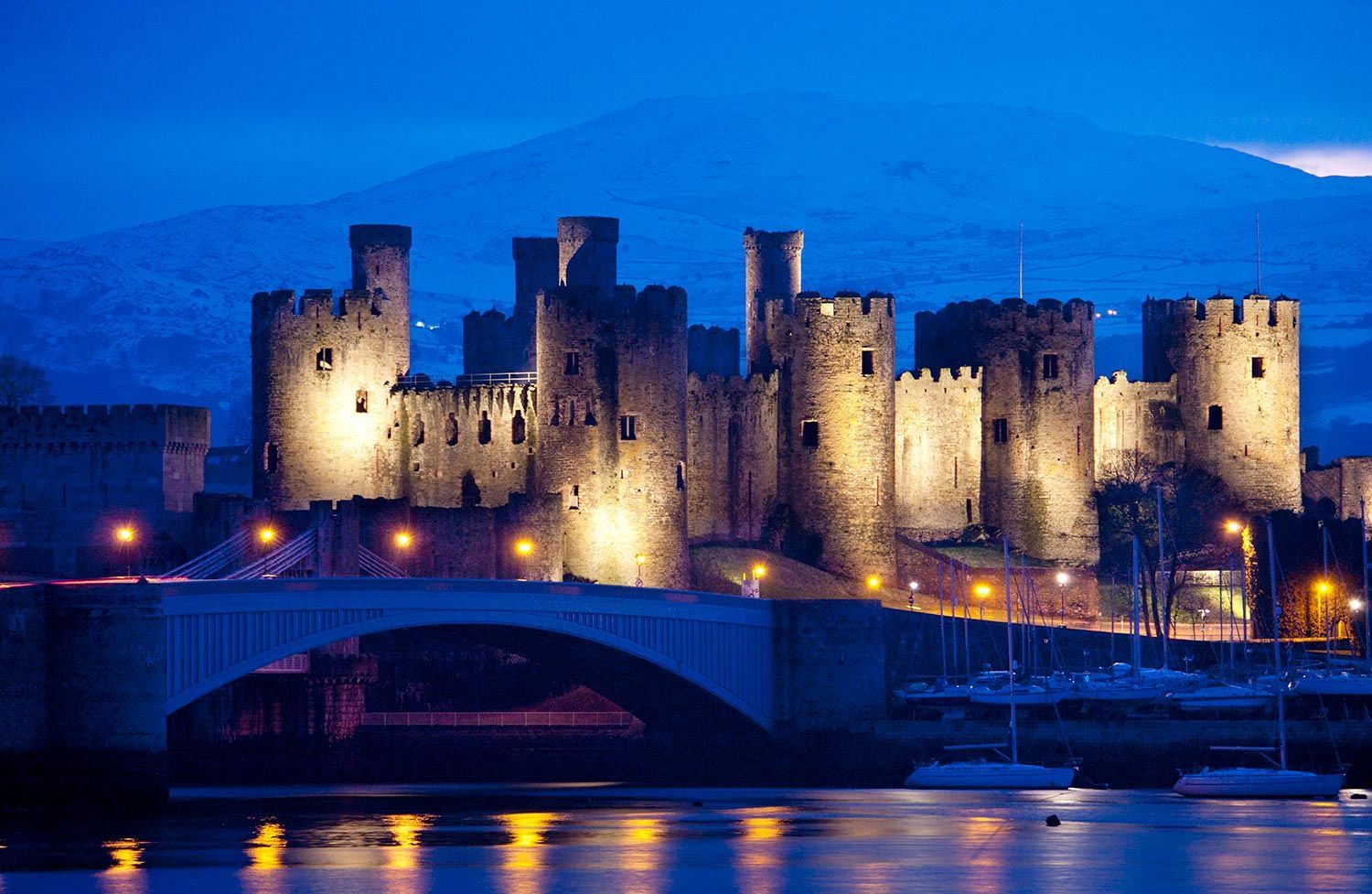 Conwy Castle, Wales, illuminated at night. Dark stone fortress with towers reflects in water below.