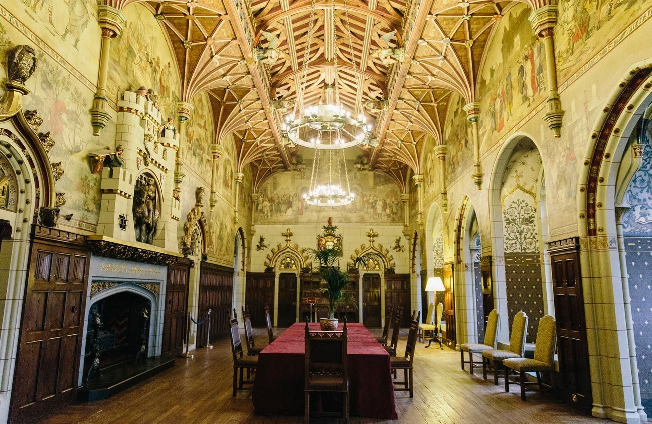 Ornate dining hall with long table, fireplace, detailed ceiling, and decorative walls.