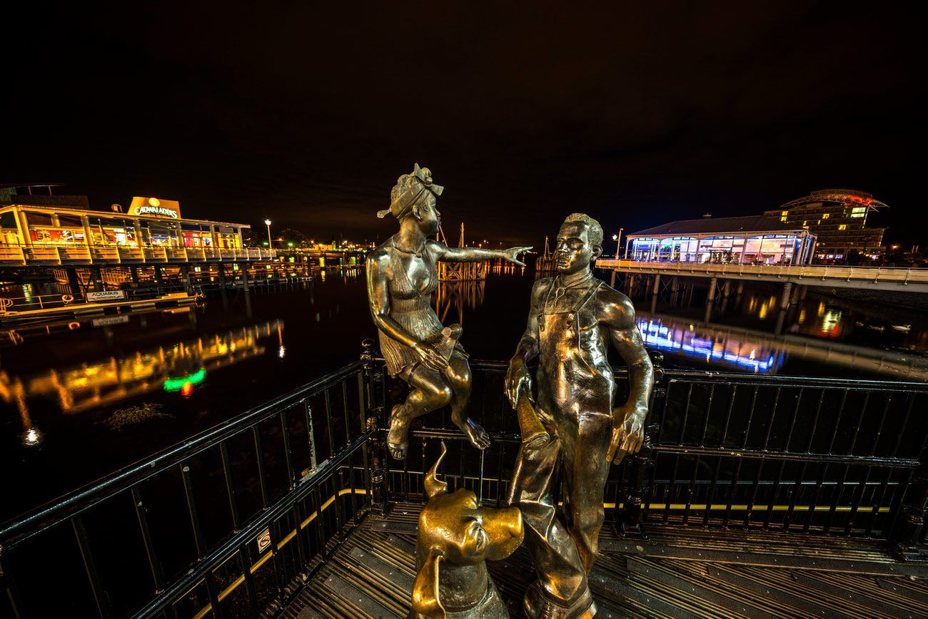Bronze statue of figures on a pier at night; lit buildings and water reflections in the background.