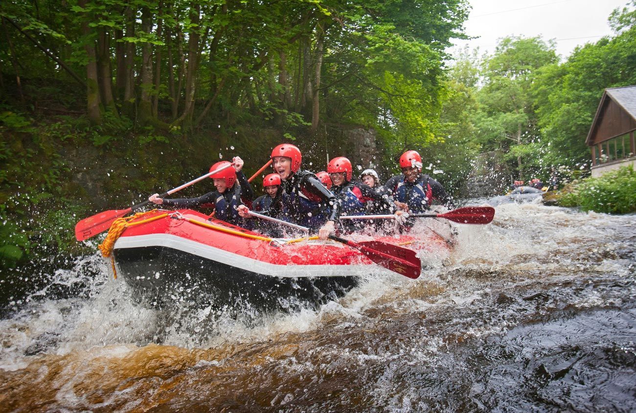People in a red raft navigate rapids, paddling with orange oars; trees line the riverbanks.