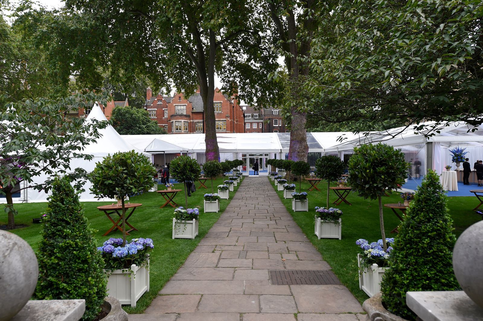 Stone path leading to white tents, lined with potted trees and flowers in a garden setting.