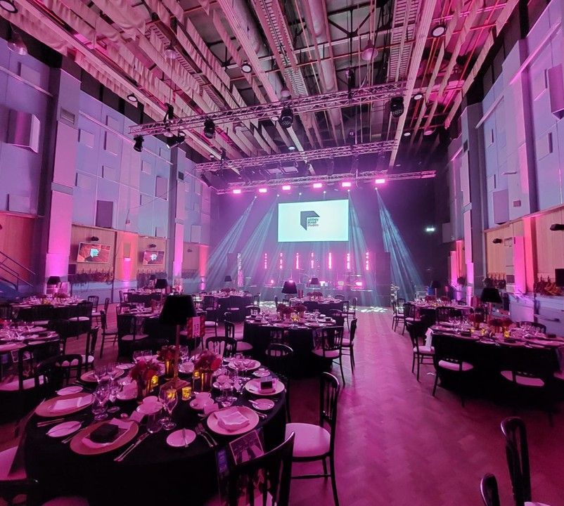 Event hall with tables set for a meal, stage with logo and lights. Pink and purple lighting.
