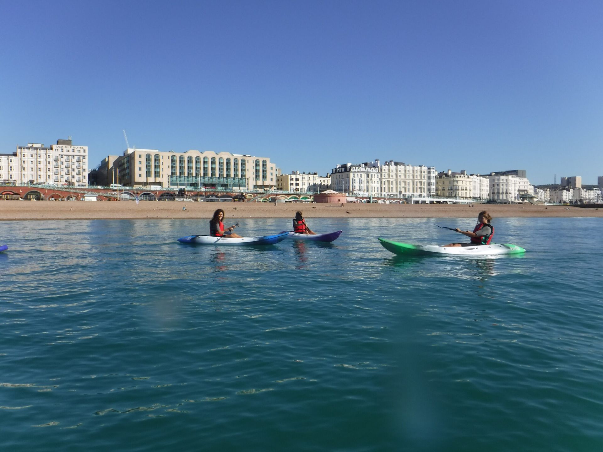 People kayaking on calm, turquoise water near a beach and buildings on a sunny day in Brighton.