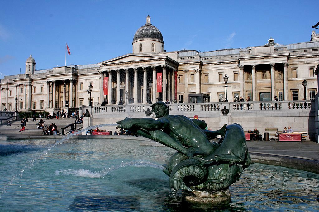 Fountain with statue in front of the National Gallery in Trafalgar Square, London, with blue sky.