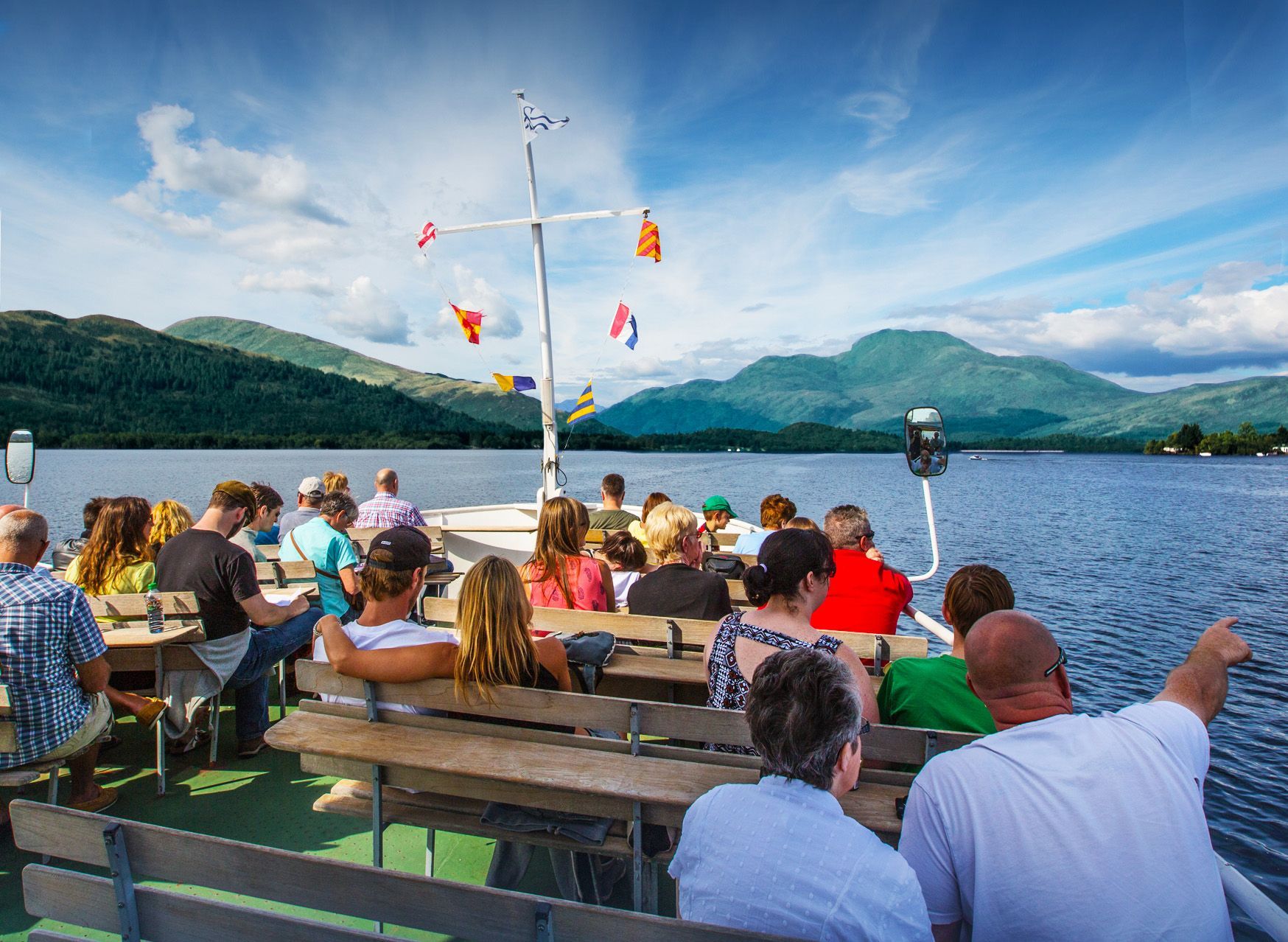 People on a boat tour on a lake with mountains in the background, blue sky, and colorful flags.