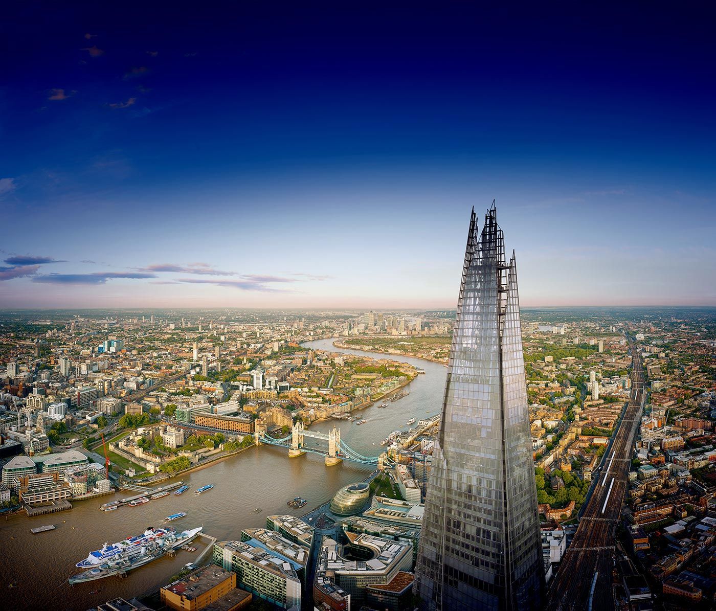The Shard skyscraper dominates London skyline, River Thames flows past Tower Bridge and cityscape below.