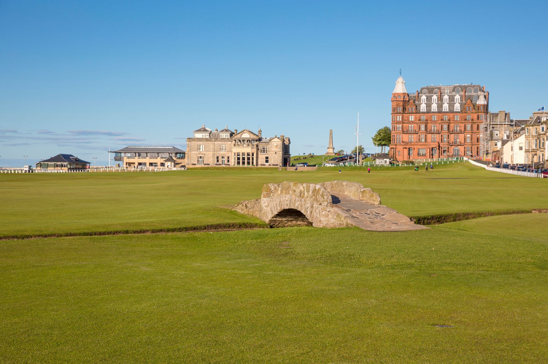 Stone bridge on a green field with buildings in the background under a blue sky.