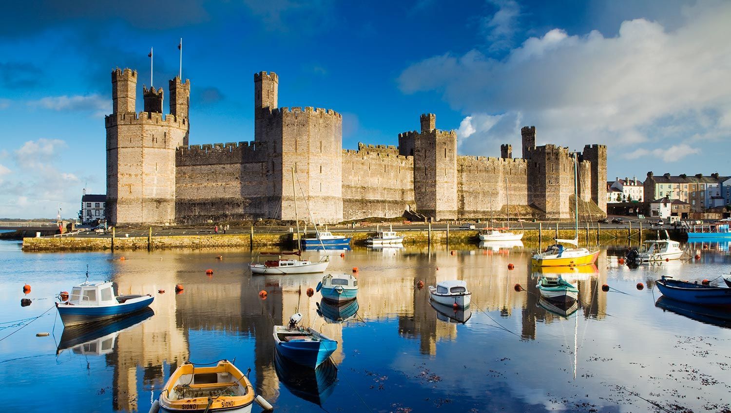 Caernarfon Castle in Wales, stone structure on waterfront with boats in calm water, blue sky.