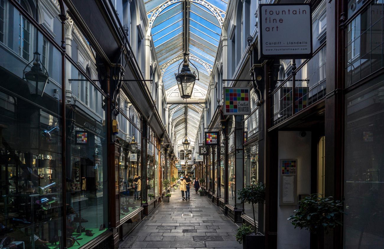 A long, glass-roofed shopping arcade with shops on both sides, and a few people walking in the distance.