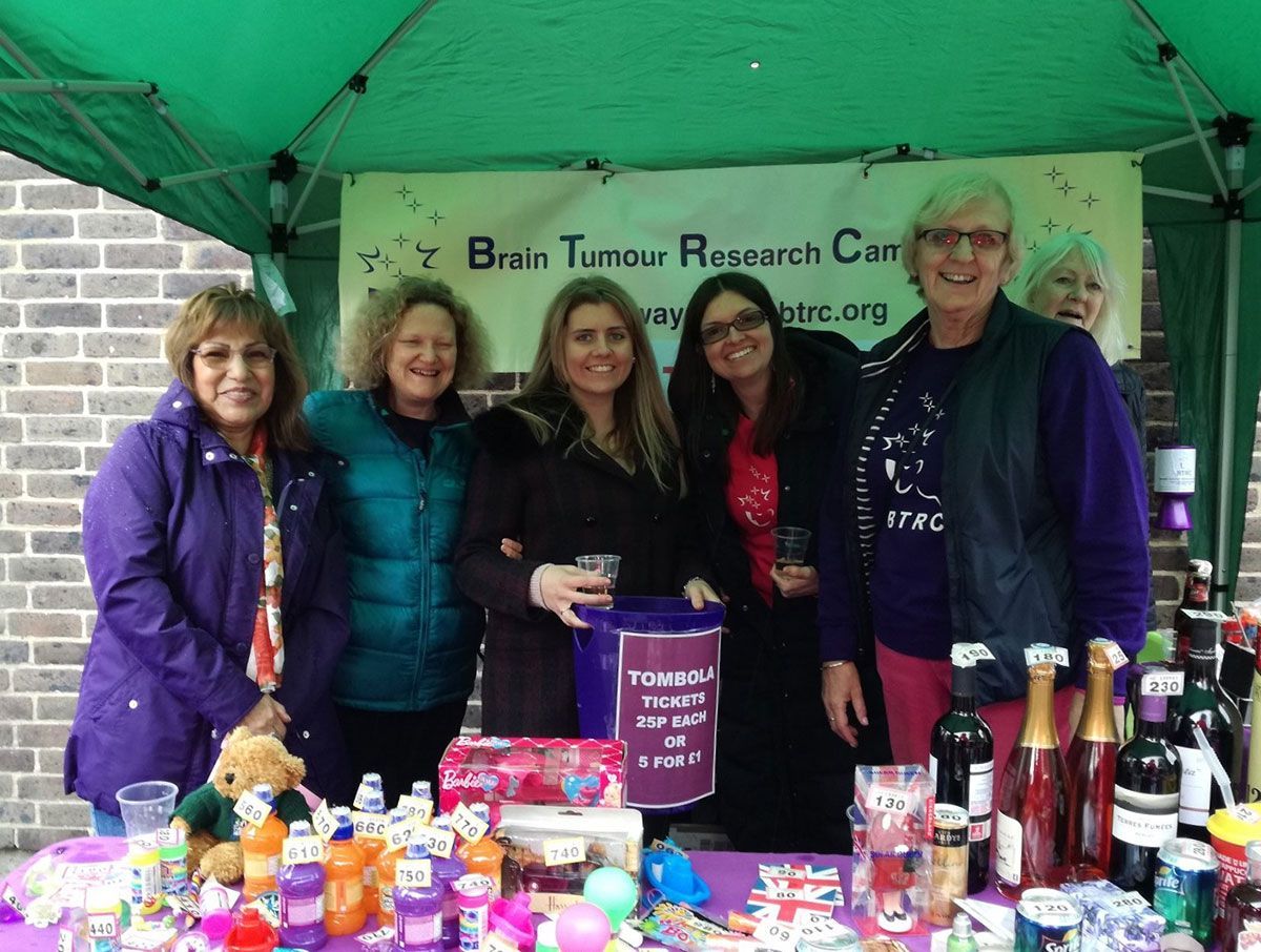 Women at a Brain Tumour Research Center fundraising stall. Smiling, selling items, under a green canopy.