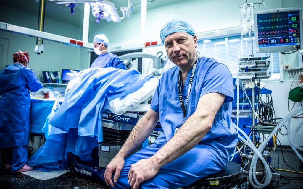 Surgeon in blue scrubs poses in operating room, other medical staff and equipment visible.
