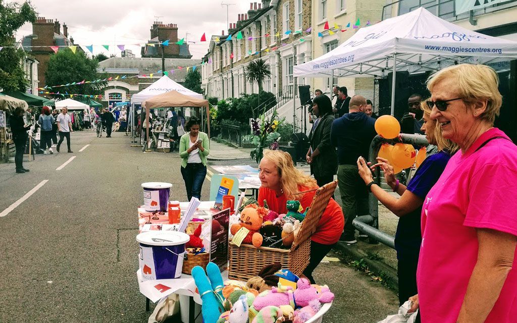 Street fair: people browsing tables with goods, tents, buildings in background, sunny day.
