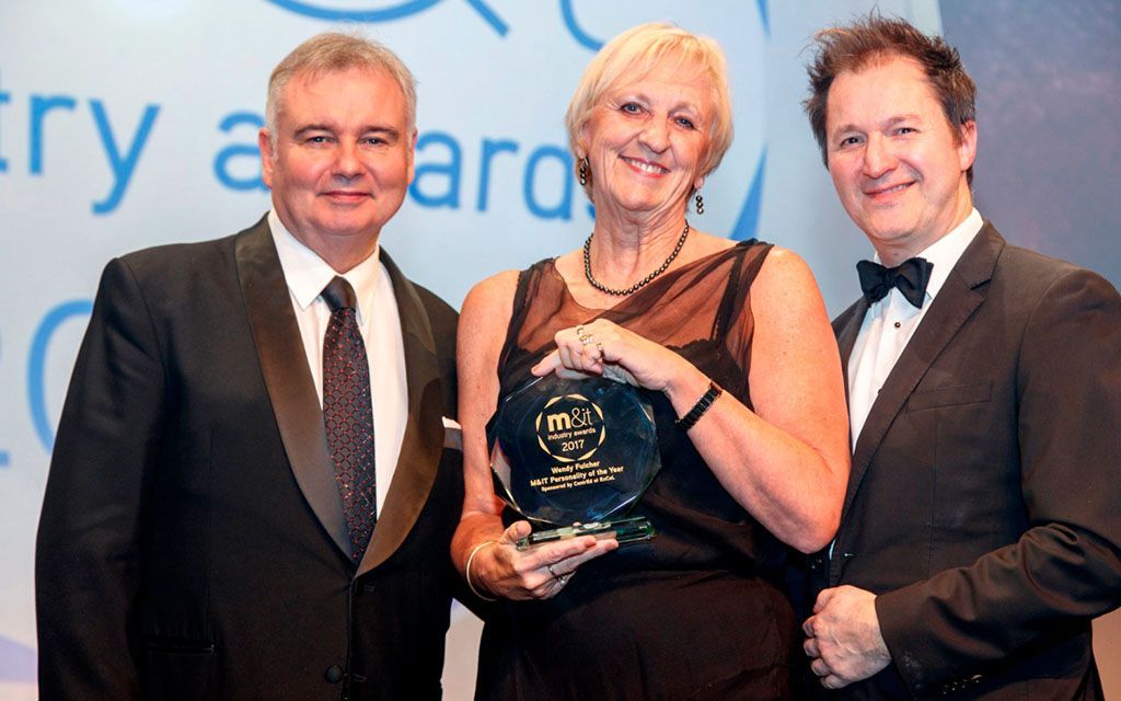 Three people at an awards ceremony; woman holding award smiles between two men in suits.