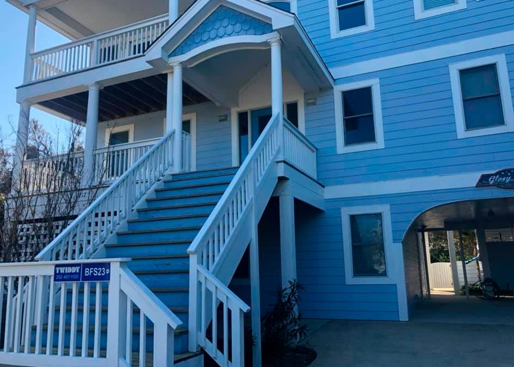 Blue beach house with stairs leading to the entrance. Open carport on the right.