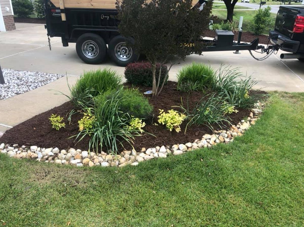 Landscaped garden bed with rocks, mulch, and various green plants in front of a lawn.