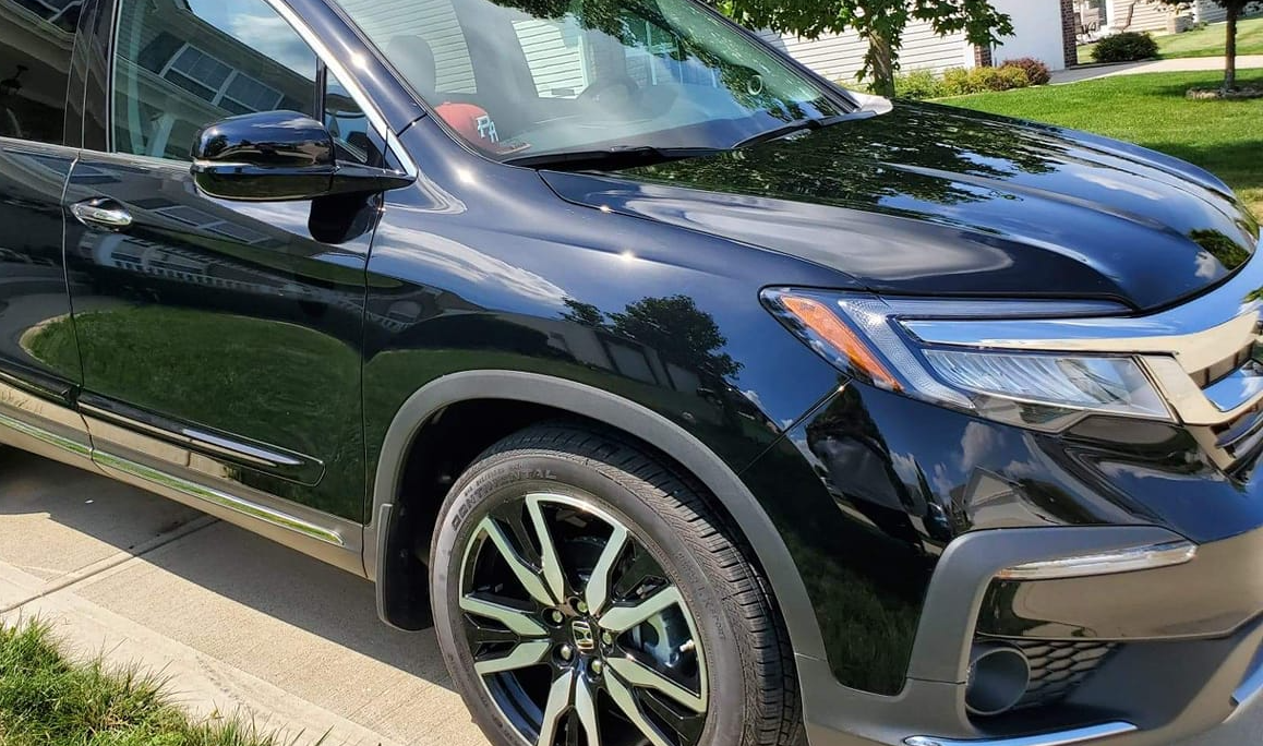 Black Honda Pilot SUV parked on a paved driveway with green grass and trees in the background.