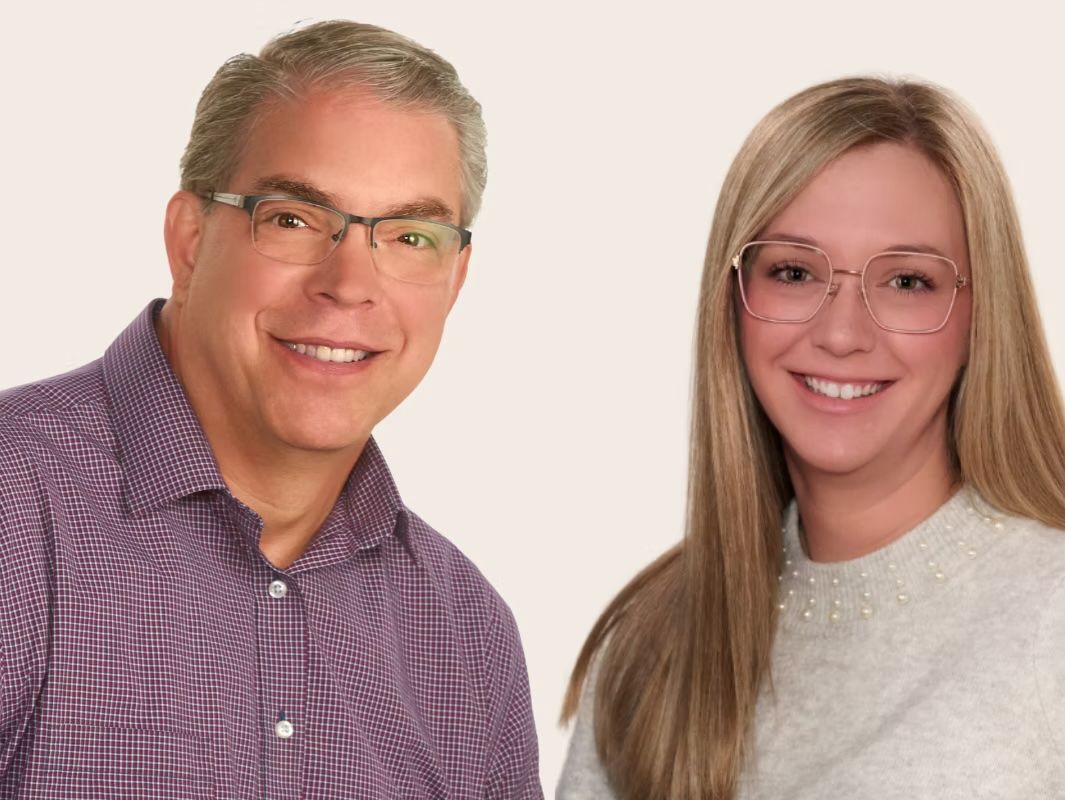Two smiling people posed against a light background, wearing glasses and casual clothing.