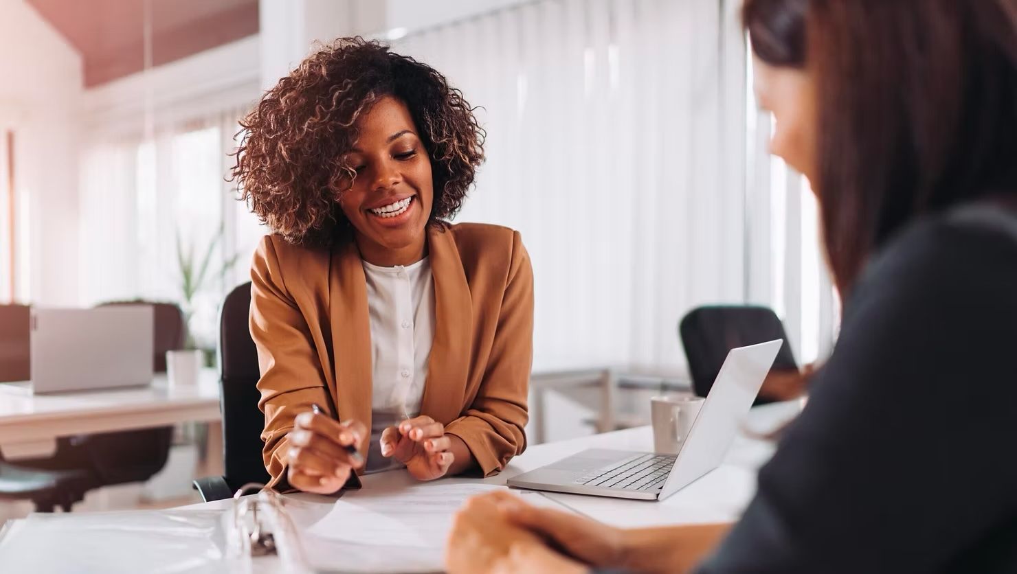 Two people in a bright office discussing documents across a desk, one smiling and holding a pen.