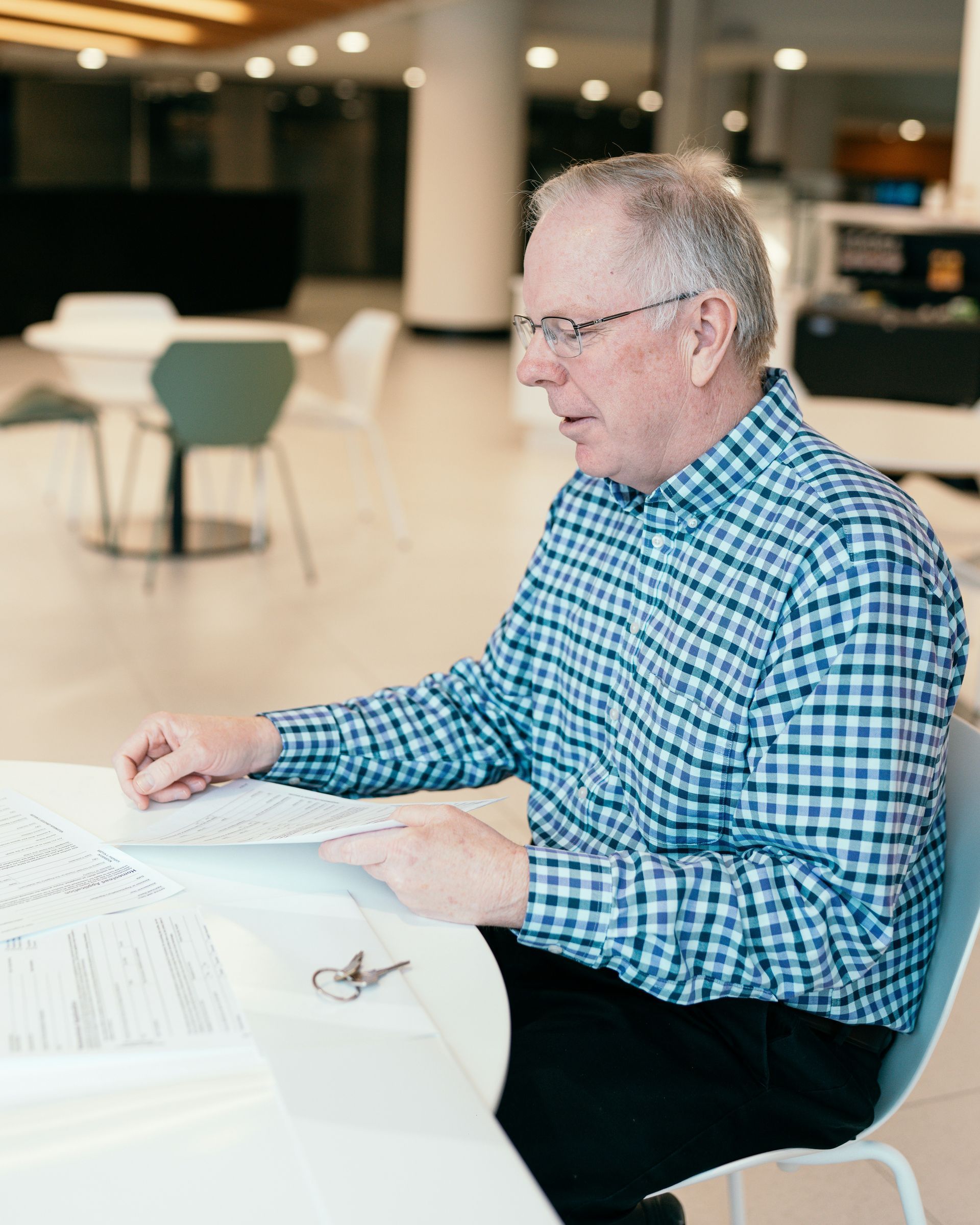 Man in a blue checkered shirt sitting at a table and reviewing papers in a bright lobby