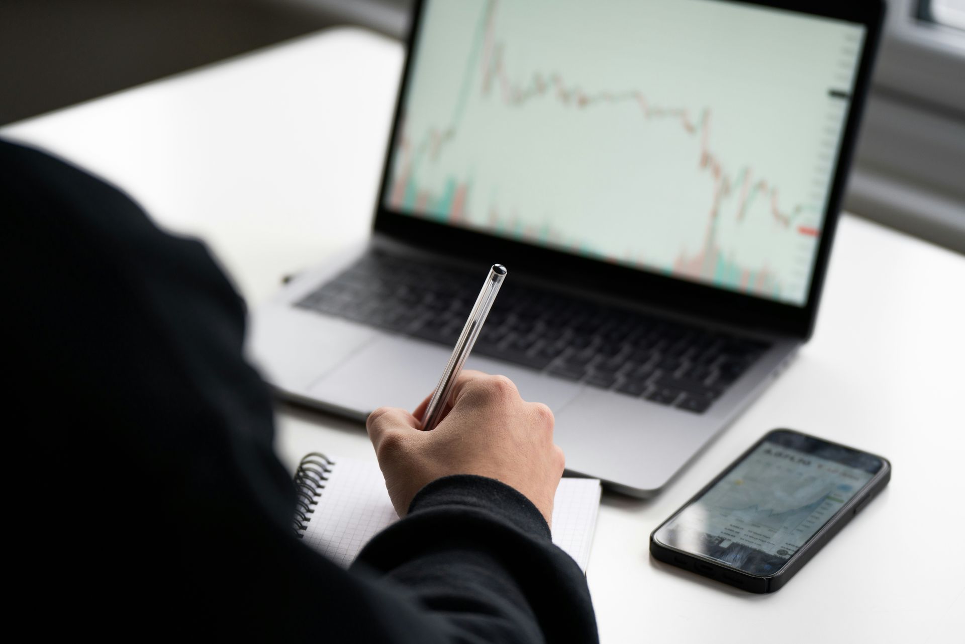 Person taking notes beside laptop with stock chart and smartphone on desk