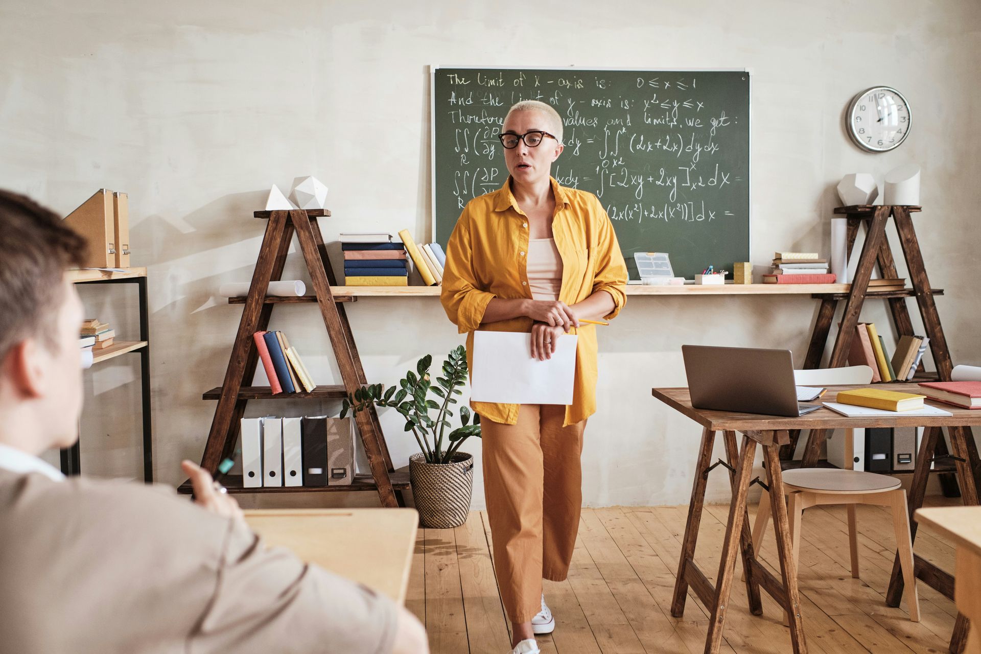 Teacher presenting in a classroom with chalkboard, bookshelves, and a laptop on a desk.