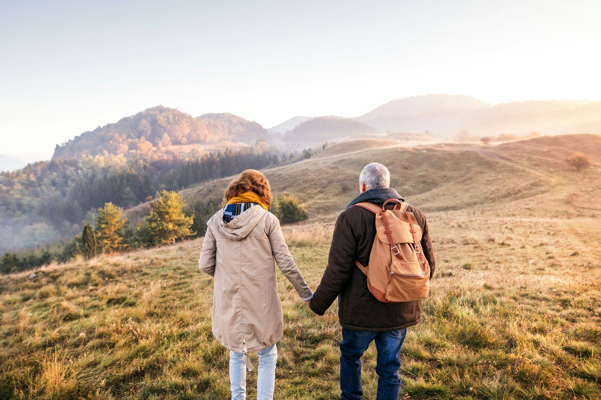 Two hikers holding hands on a grassy hill overlooking misty mountains at sunrise