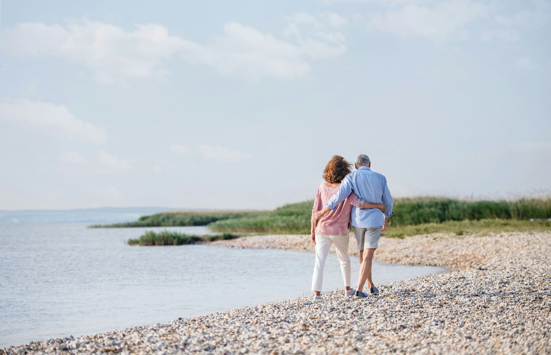 Two people walking hand in hand along a rocky beach by calm water and grass under a blue sky