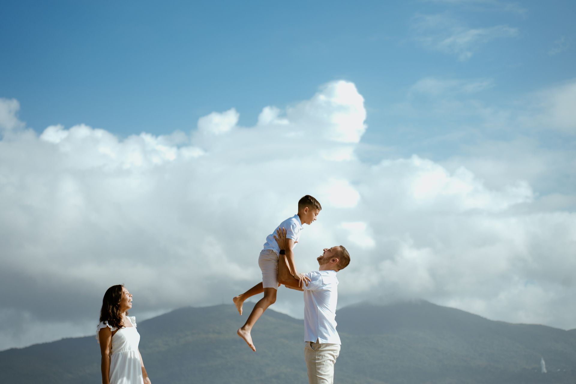 Man lifting a child against a bright cloudy sky, with a woman standing nearby and mountains in the distance