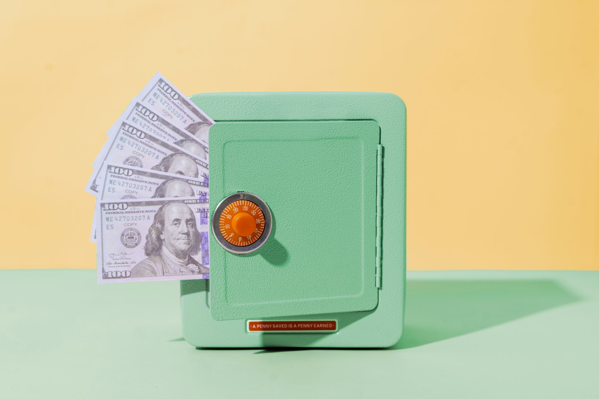 Mint green safe with orange dial, partly open with paper bills sticking out on a pastel background