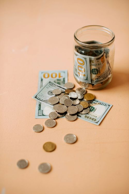 Glass jar and scattered coins on a peach surface, with folded cash nearby.