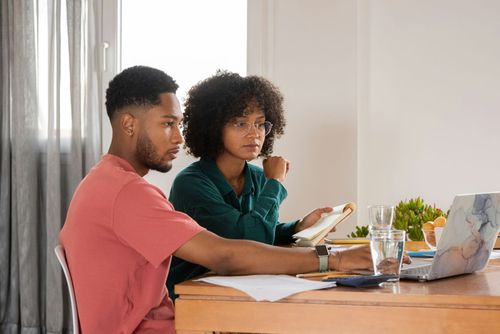 Two people working at a table with a laptop, papers, and a notebook in a bright office setting