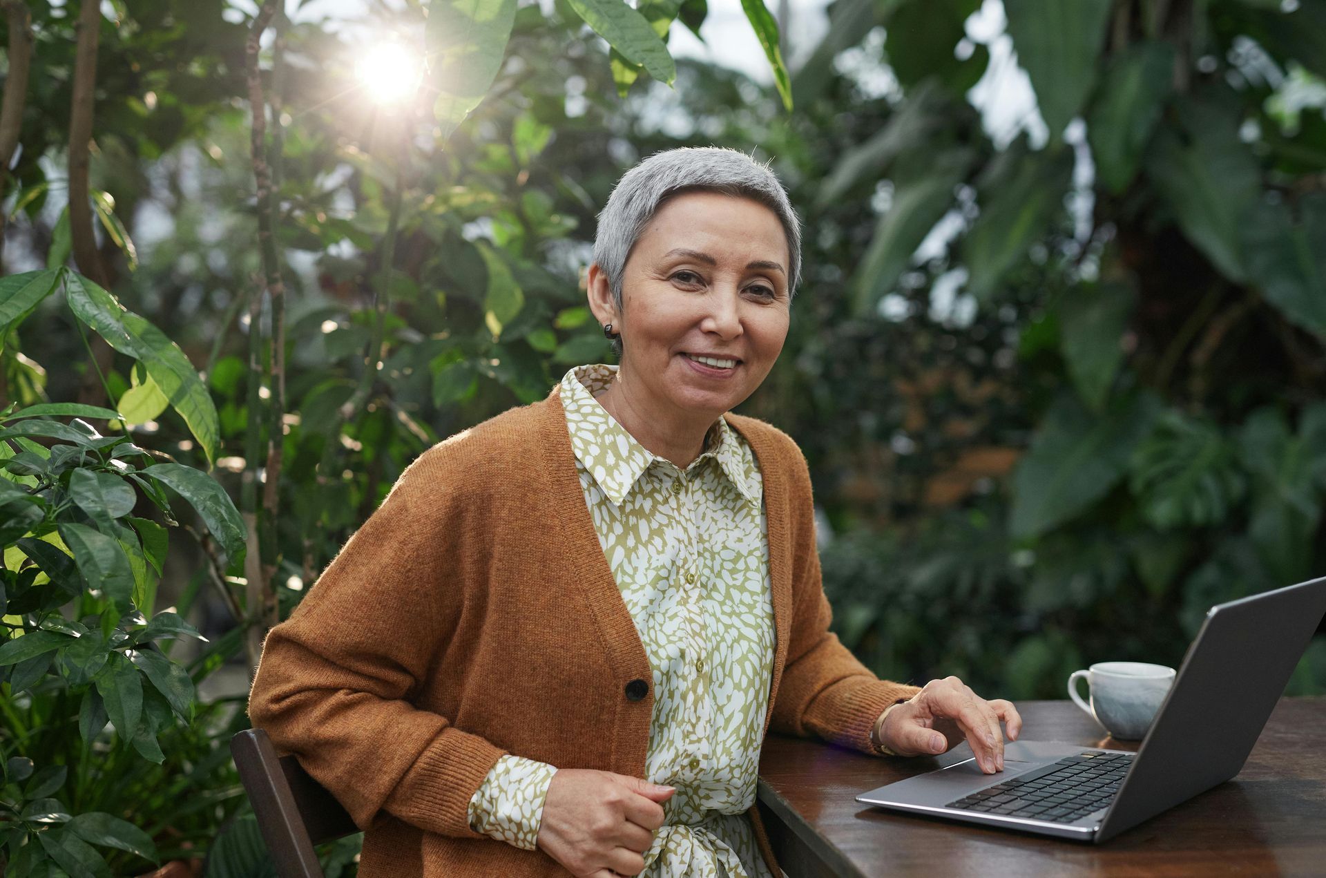Woman smiling beside a laptop at an outdoor table in a lush garden