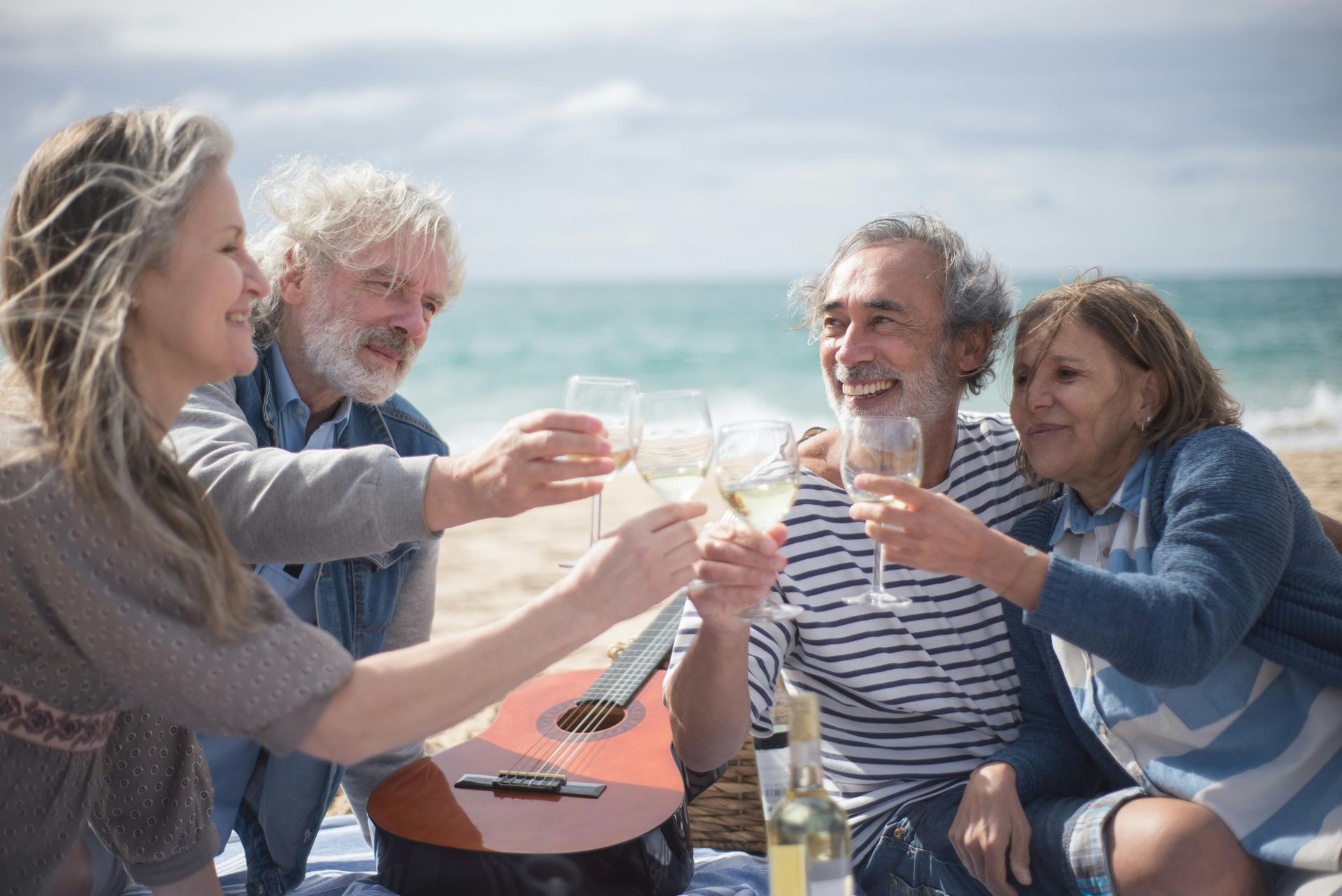 Four friends toast drinks at a seaside picnic table on a sunny day