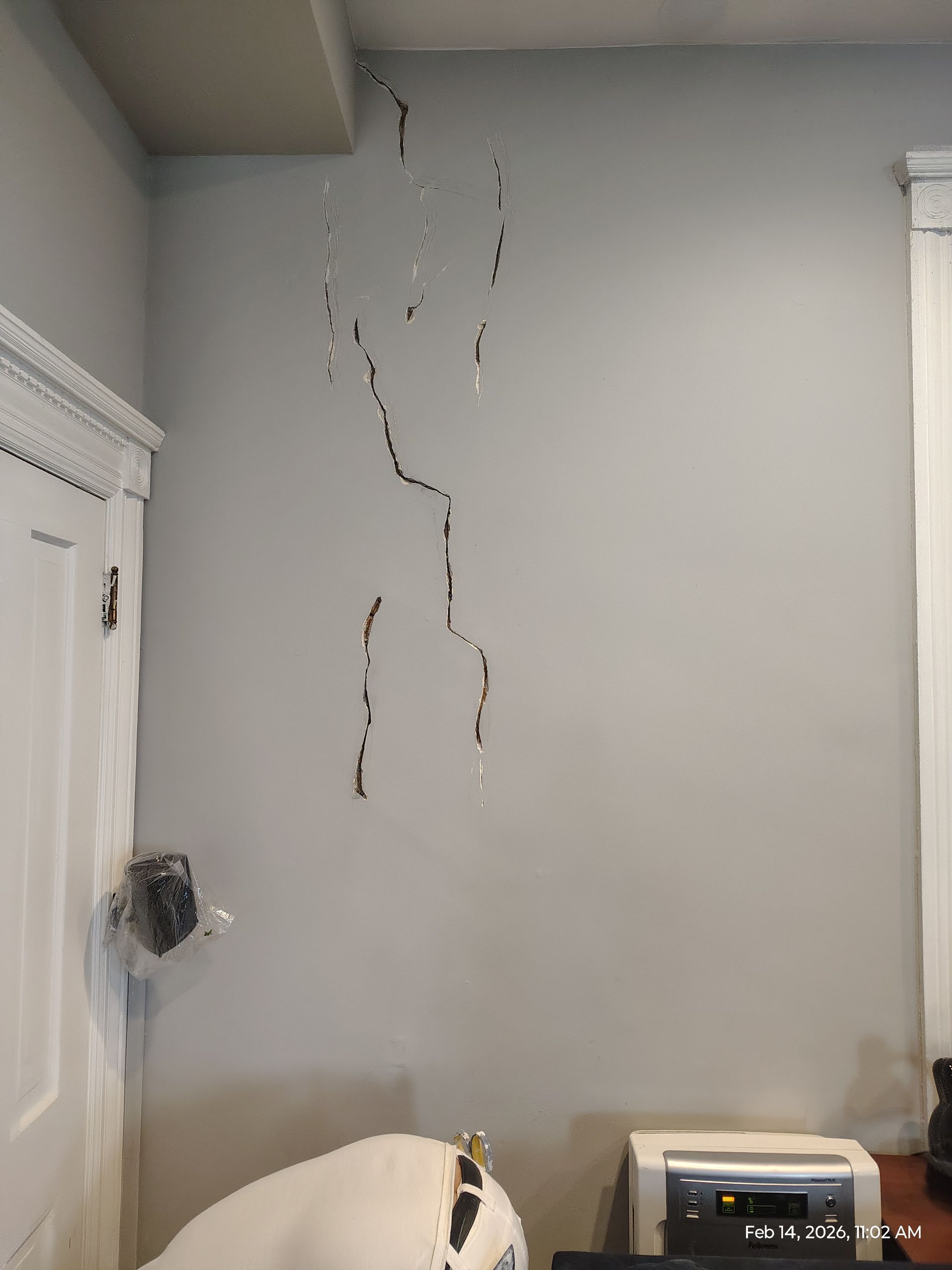 White wall with long vertical cracks and peeling damage above a partially visible bed and printer in a room