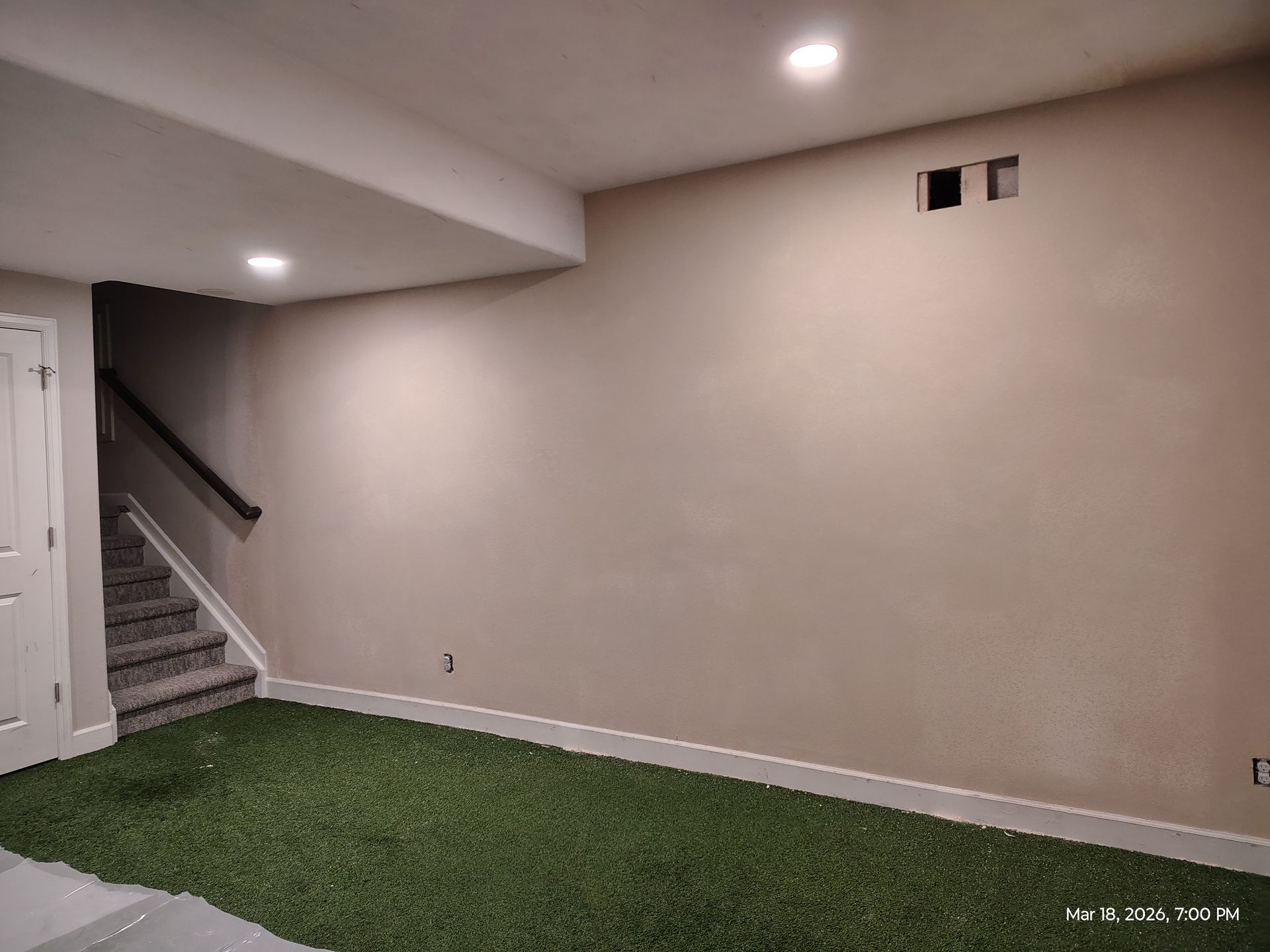 Basement room with beige walls, stairway on left, and green carpeted floor.