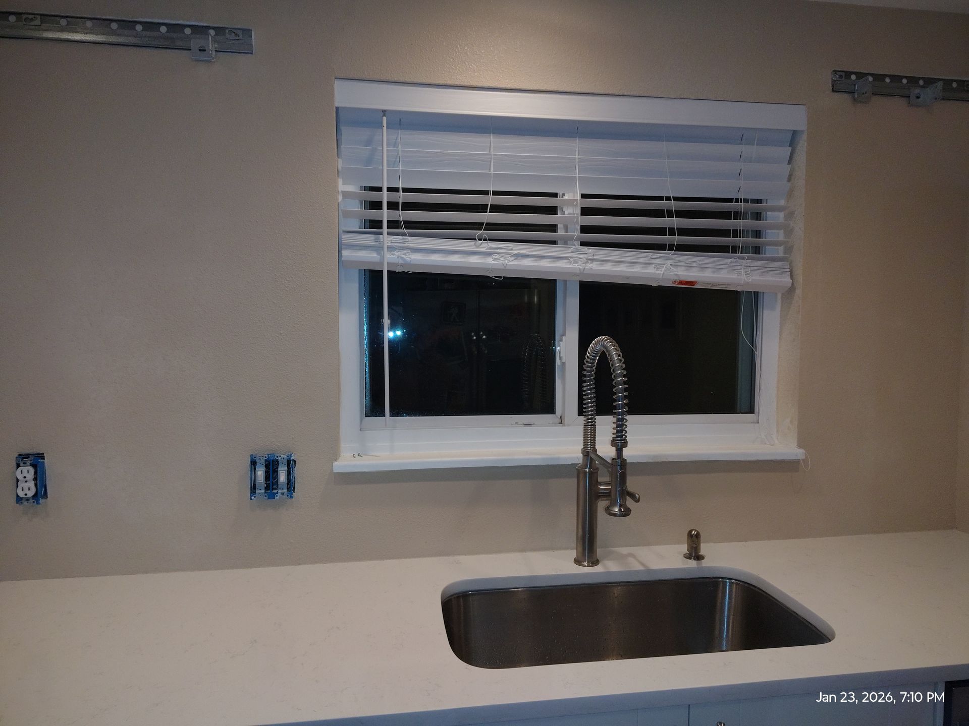 Empty kitchen with a window above a stainless steel sink and faucet, beige walls, and unfinished outlets.