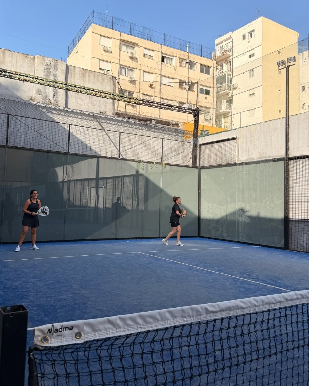 Dos personas practicando en una cancha de tenis azul al aire libre rodeada de edificios altos.