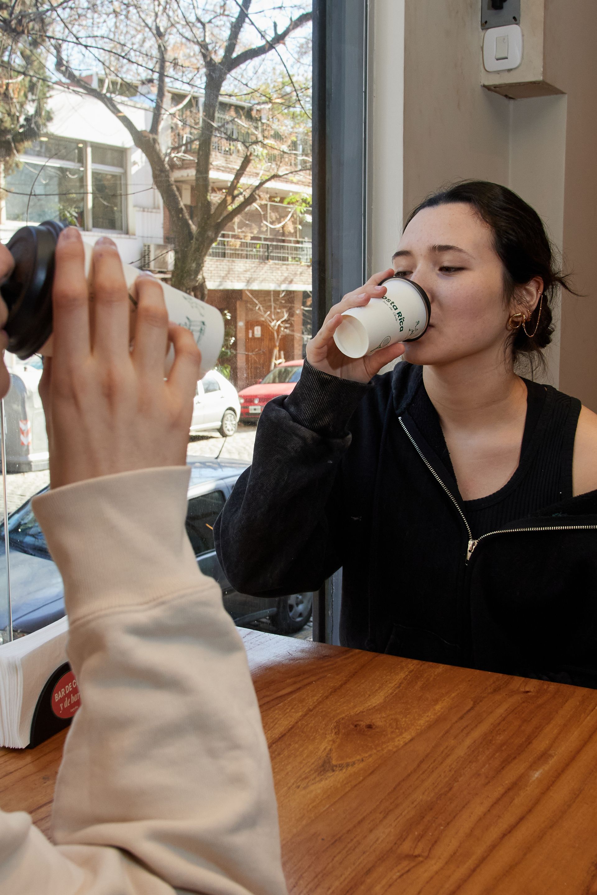 Dos personas sentadas en una mesa de un café; una toma una foto mientras la otra bebe de una taza junto a la ventana.